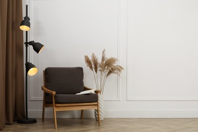 Stylish armchair with book, floor lamp and spikes near white wall indoors, space for text. Interior design