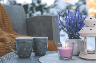 Photo of Burning candle, lantern, potted flowers and cups with hot drink on coffee table outdoors. Cosy winter