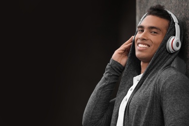 Photo of Handsome young African-American man with headphones listening to music near wall on city street. Space for text