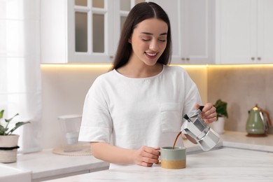 Photo of Happy woman pouring coffee from jezve into cup at white marble table in kitchen. Lazy morning
