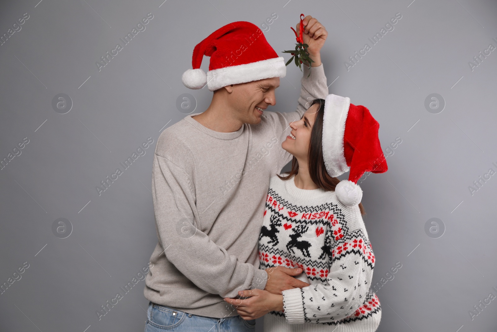 Photo of Happy couple in Santa hats standing under mistletoe bunch on grey background