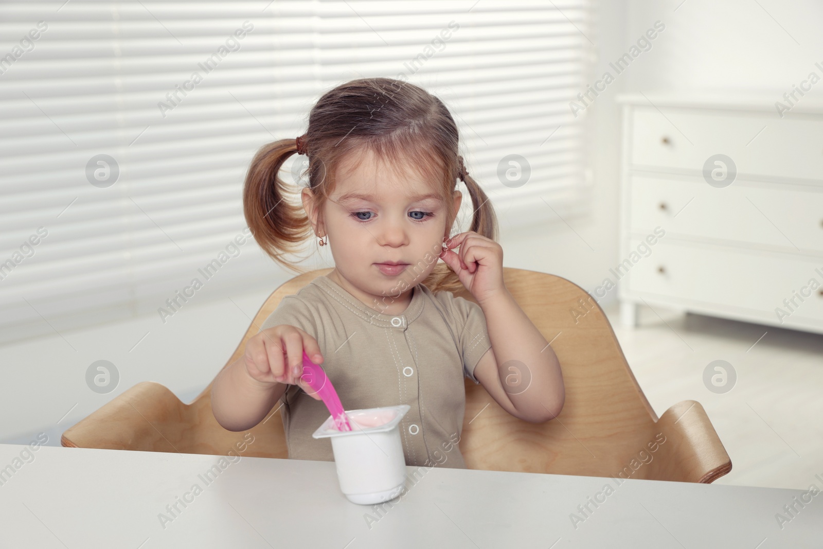 Photo of Cute little child eating tasty yogurt from plastic cup with spoon at white table indoors