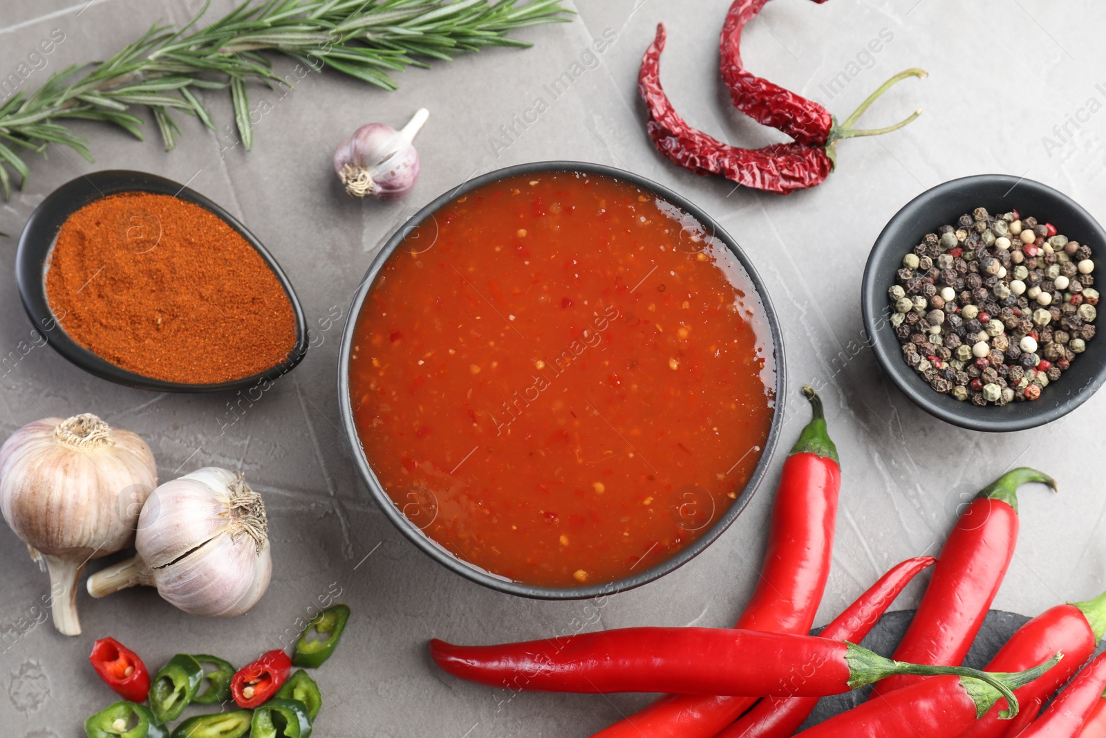 Photo of Spicy chili sauce in bowl and ingredients on grey table, flat lay