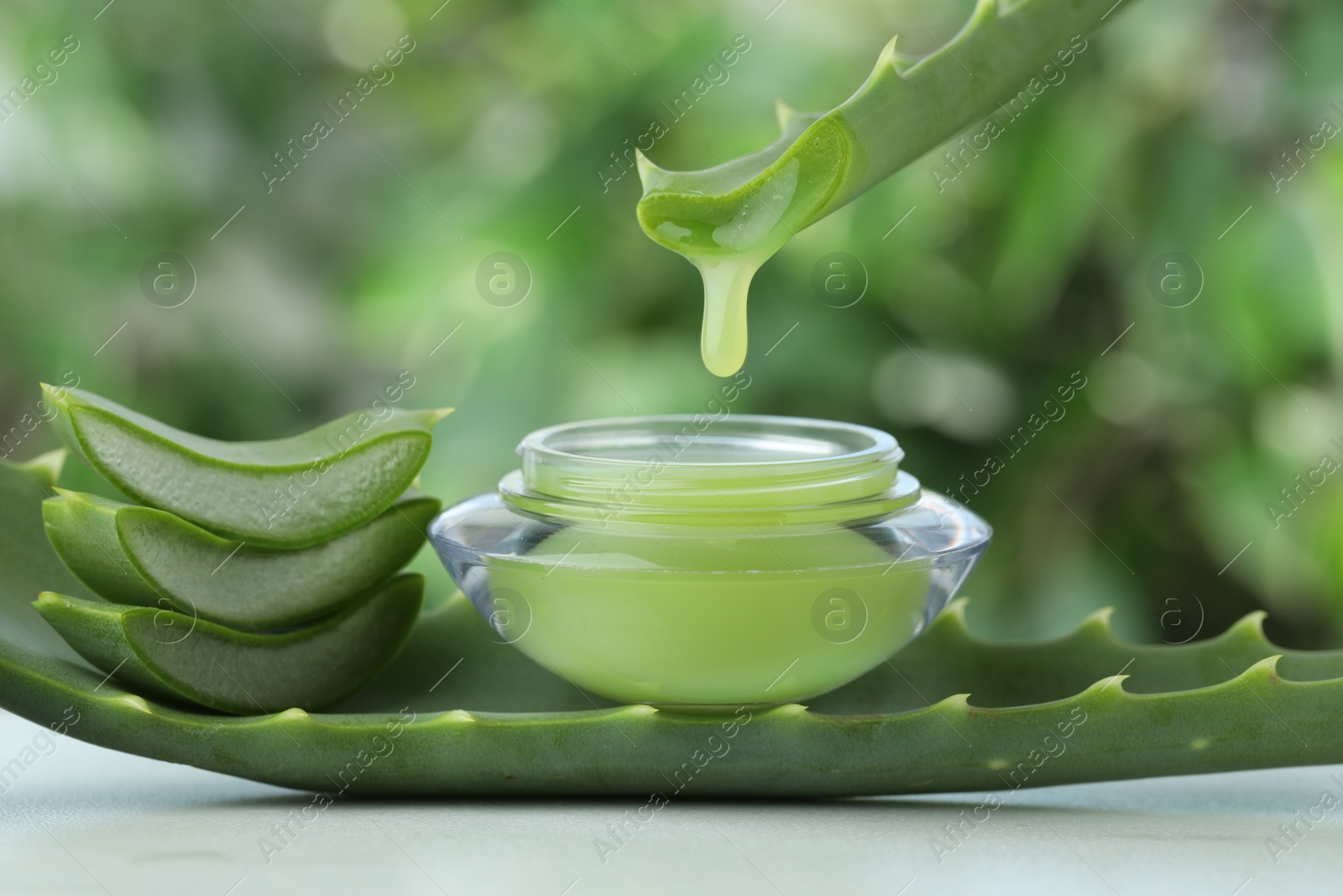 Photo of Aloe vera juice dripping from leaf into jar with cream on white table against green blurred background, closeup