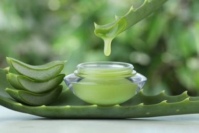 Aloe vera juice dripping from leaf into jar with cream on white table against green blurred background, closeup