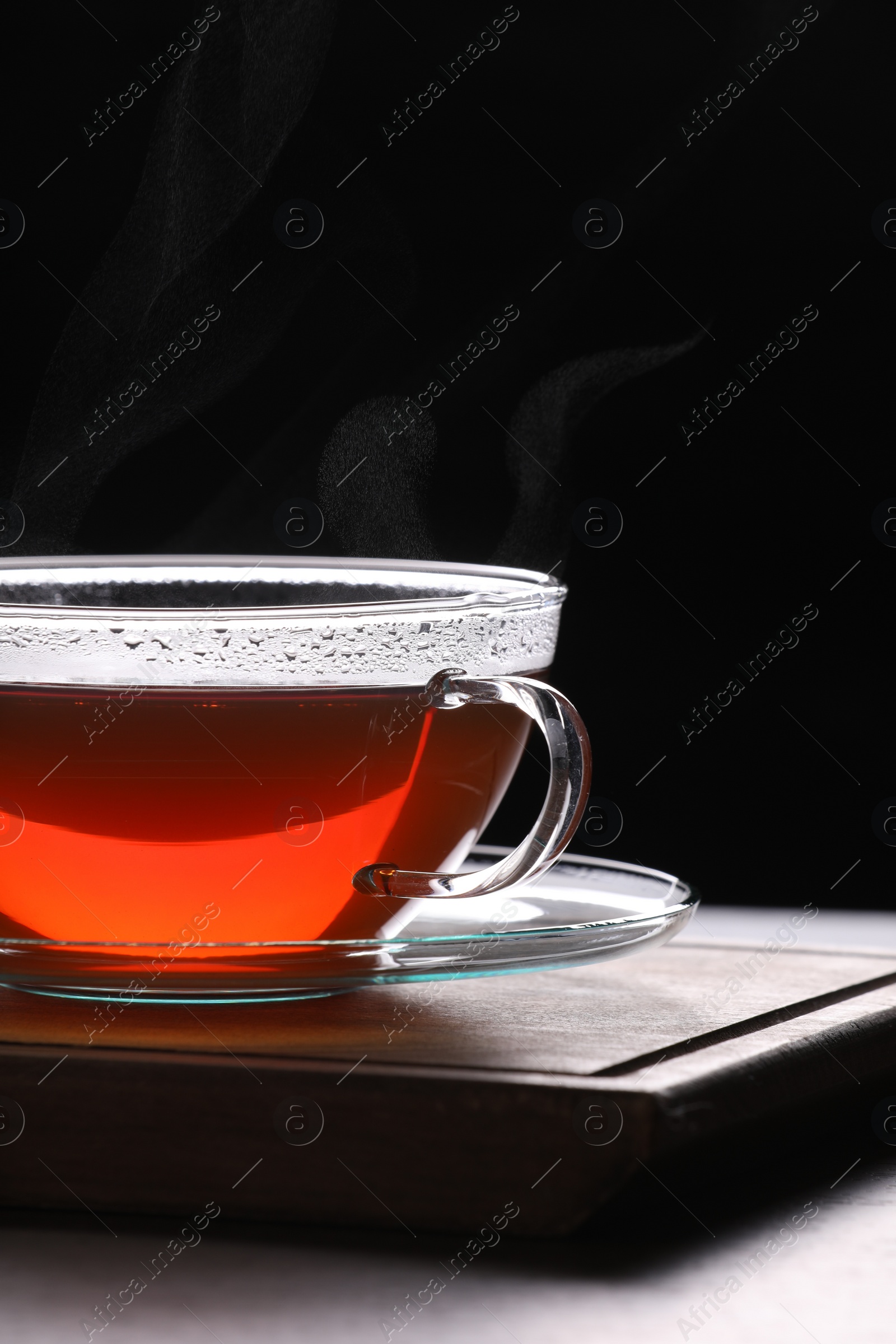 Photo of Glass cup of tea with saucer on table against black background