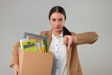 Photo of Unemployment problem. Unhappy woman with box of personal office belongings showing thumbs down on grey background