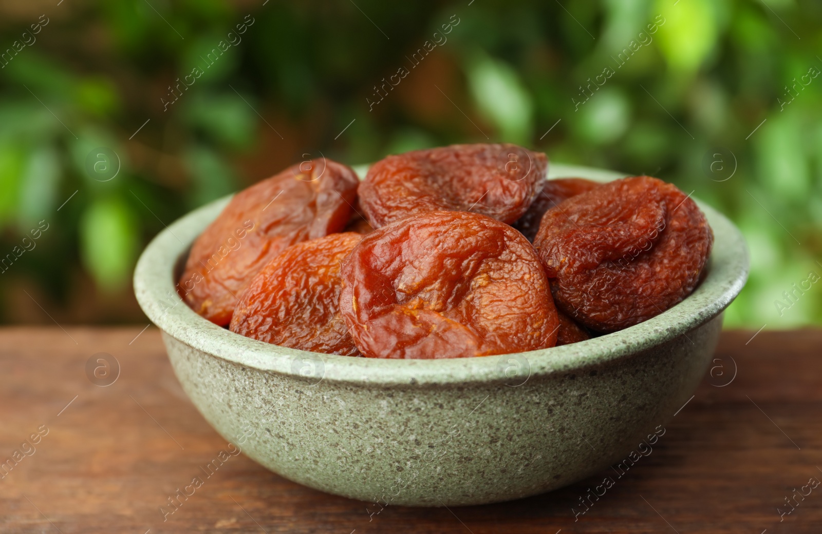 Photo of Bowl of tasty apricots on wooden table against blurred green background. Dried fruits