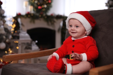 Photo of Cute little baby wearing Santa Claus suit sitting in armchair at home. Christmas celebration