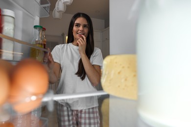 Thoughtful young woman near modern refrigerator in kitchen at night, view from inside