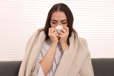 Sick woman wrapped in blanket with tissue blowing nose on sofa indoors. Cold symptoms