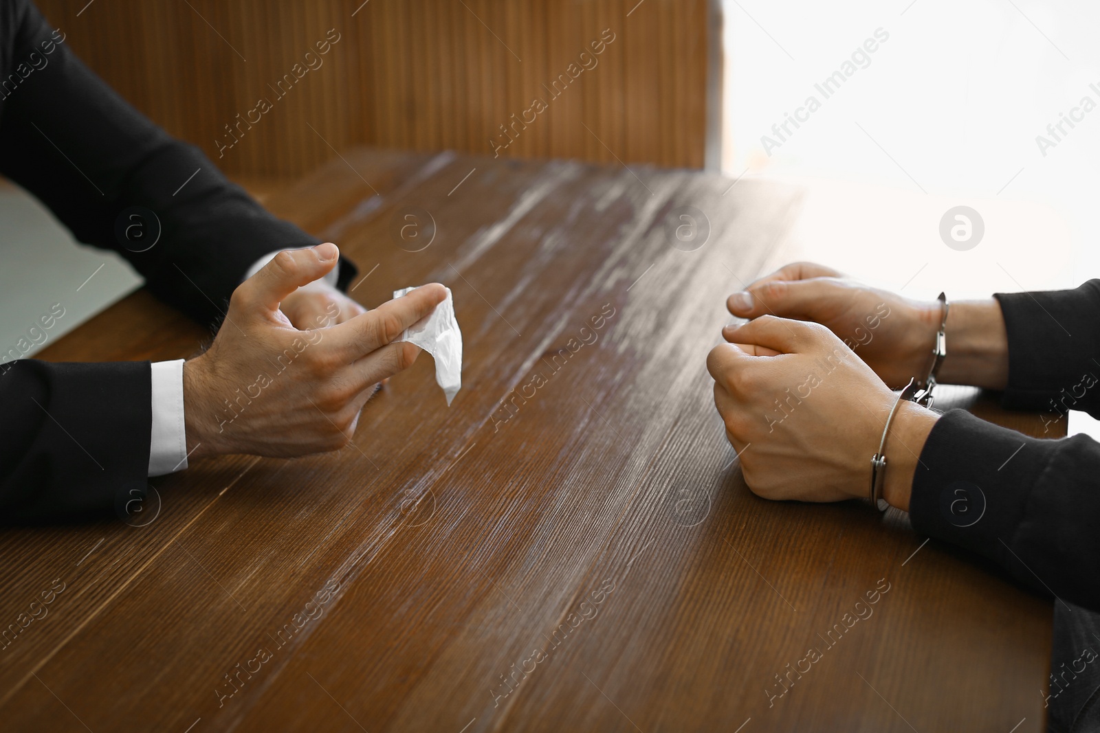 Photo of Police officer interrogating drug dealer in handcuffs at desk indoors. Criminal law