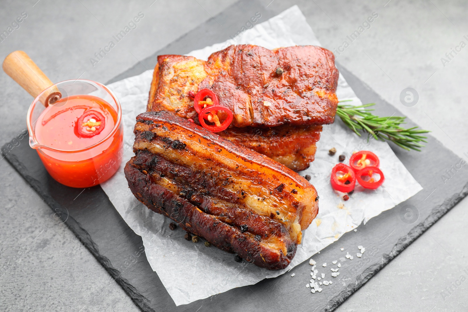 Photo of Pieces of baked pork belly served with sauce, rosemary and chili pepper on grey table