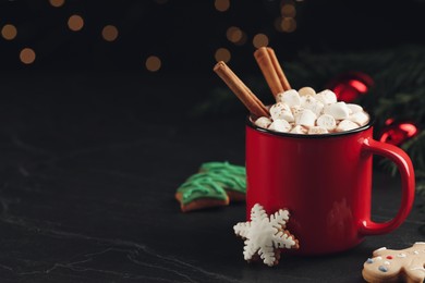Photo of Delicious hot chocolate with marshmallows, cinnamon and gingerbread cookies on black table against blurred lights, space for text