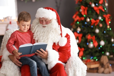 Photo of Little boy reading book while sitting on authentic Santa Claus' lap indoors