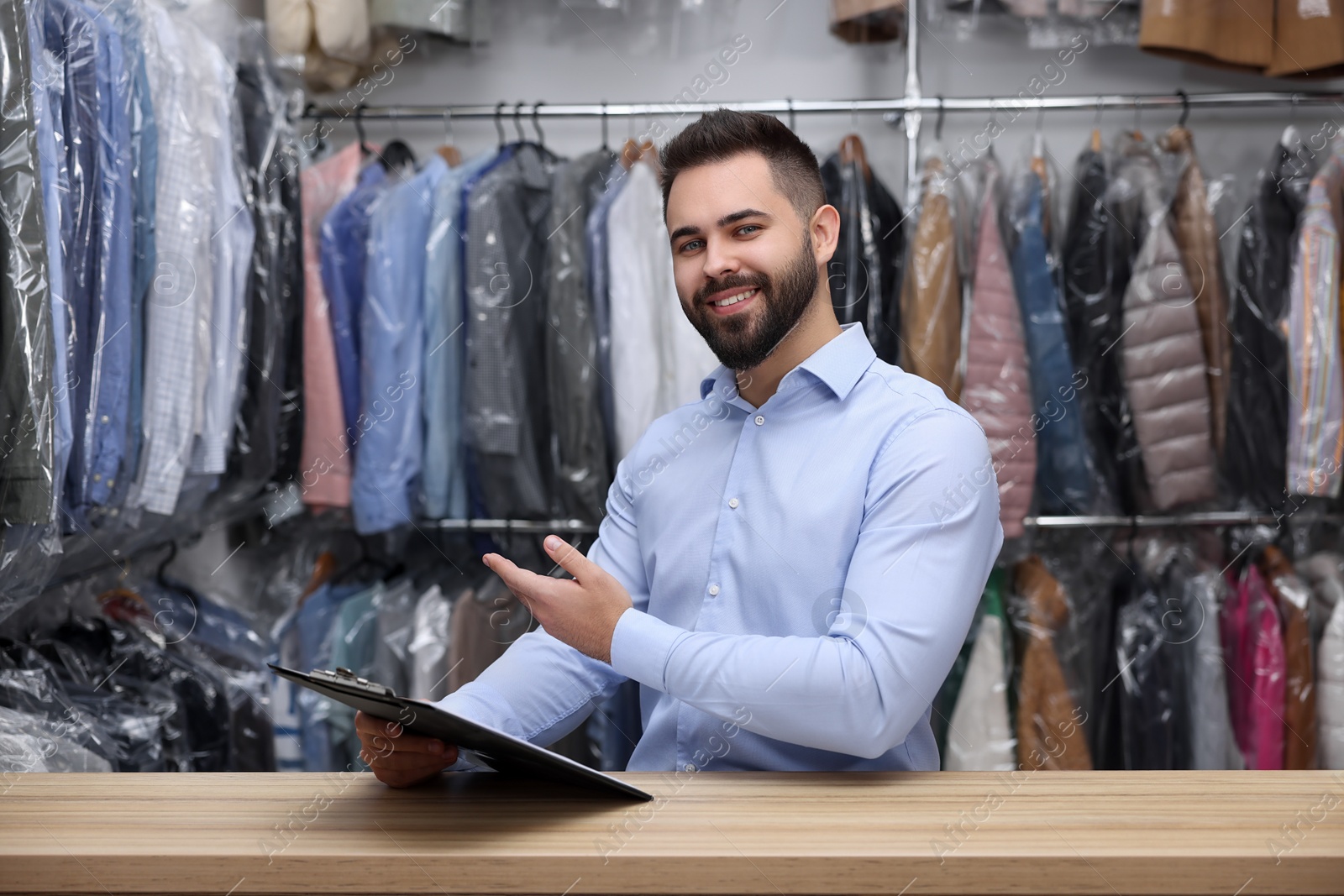 Photo of Dry-cleaning service. Happy worker holding clipboard at counter indoors