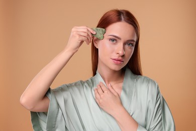 Photo of Young woman massaging her face with jade gua sha tool on pale orange background