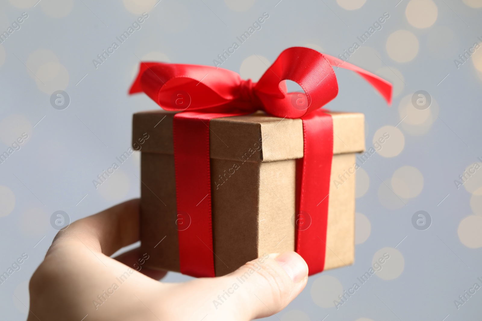 Photo of Woman holding gift box with red bow against blurred festive lights, closeup. Bokeh effect