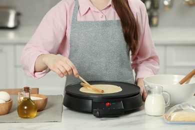 Photo of Woman cooking delicious crepe on electric maker at white marble table in kitchen, closeup