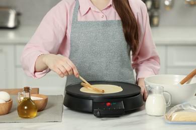 Woman cooking delicious crepe on electric maker at white marble table in kitchen, closeup