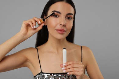 Beautiful young woman applying mascara on grey background