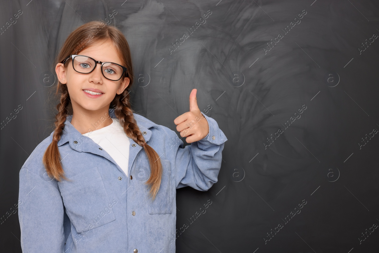 Photo of Cute schoolgirl in glasses showing thumb up near blackboard. Space for text