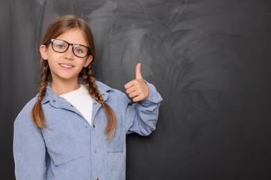 Photo of Cute schoolgirl in glasses showing thumb up near blackboard. Space for text