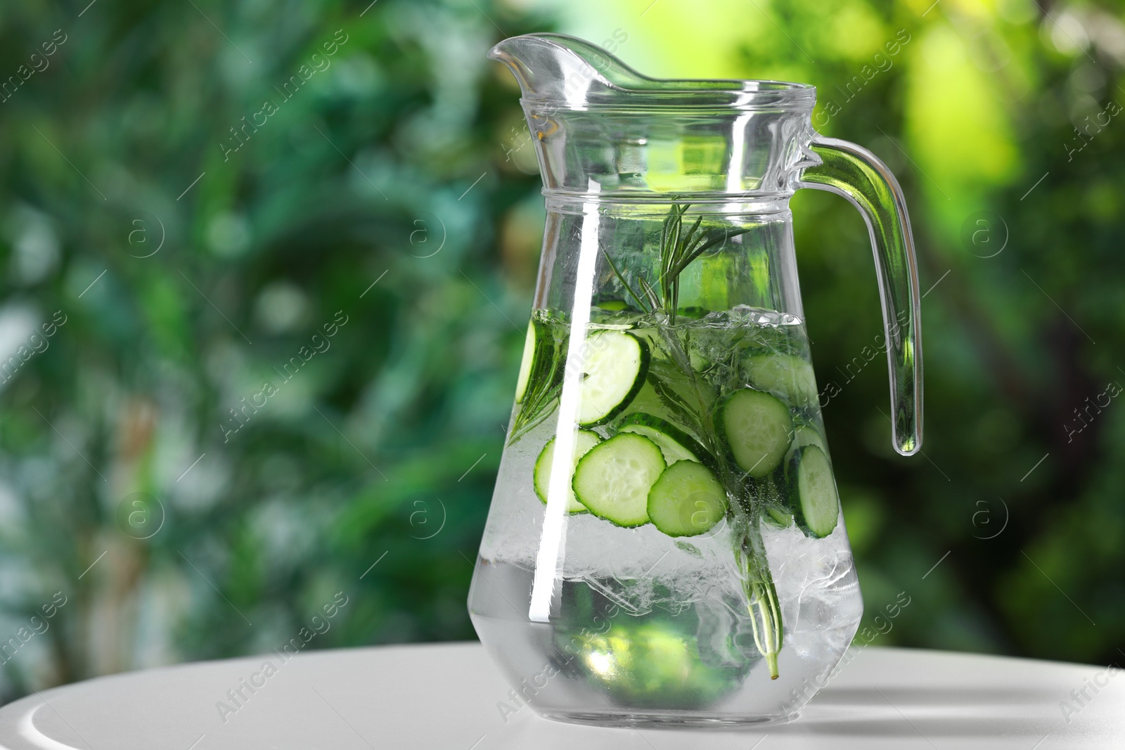 Photo of Refreshing cucumber water with rosemary in jug on white table against blurred green background, closeup. Space for text