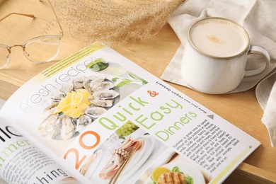 Photo of Open cooking magazine with wooden tray and cup of coffee on table, closeup