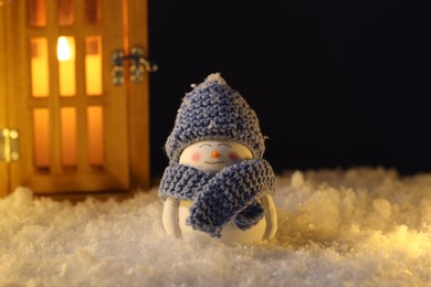 Photo of Cute decorative snowman and lantern on artificial snow against dark background