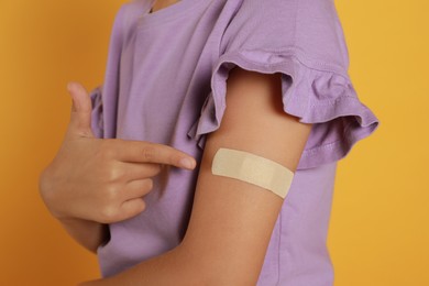 Photo of Vaccinated little girl showing medical plaster on her arm against yellow background, closeup
