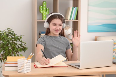 Photo of Cute girl using laptop and headphones at desk in room. Home workplace