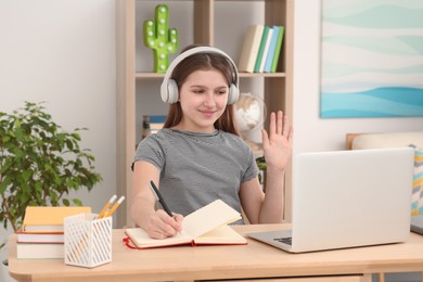 Cute girl using laptop and headphones at desk in room. Home workplace