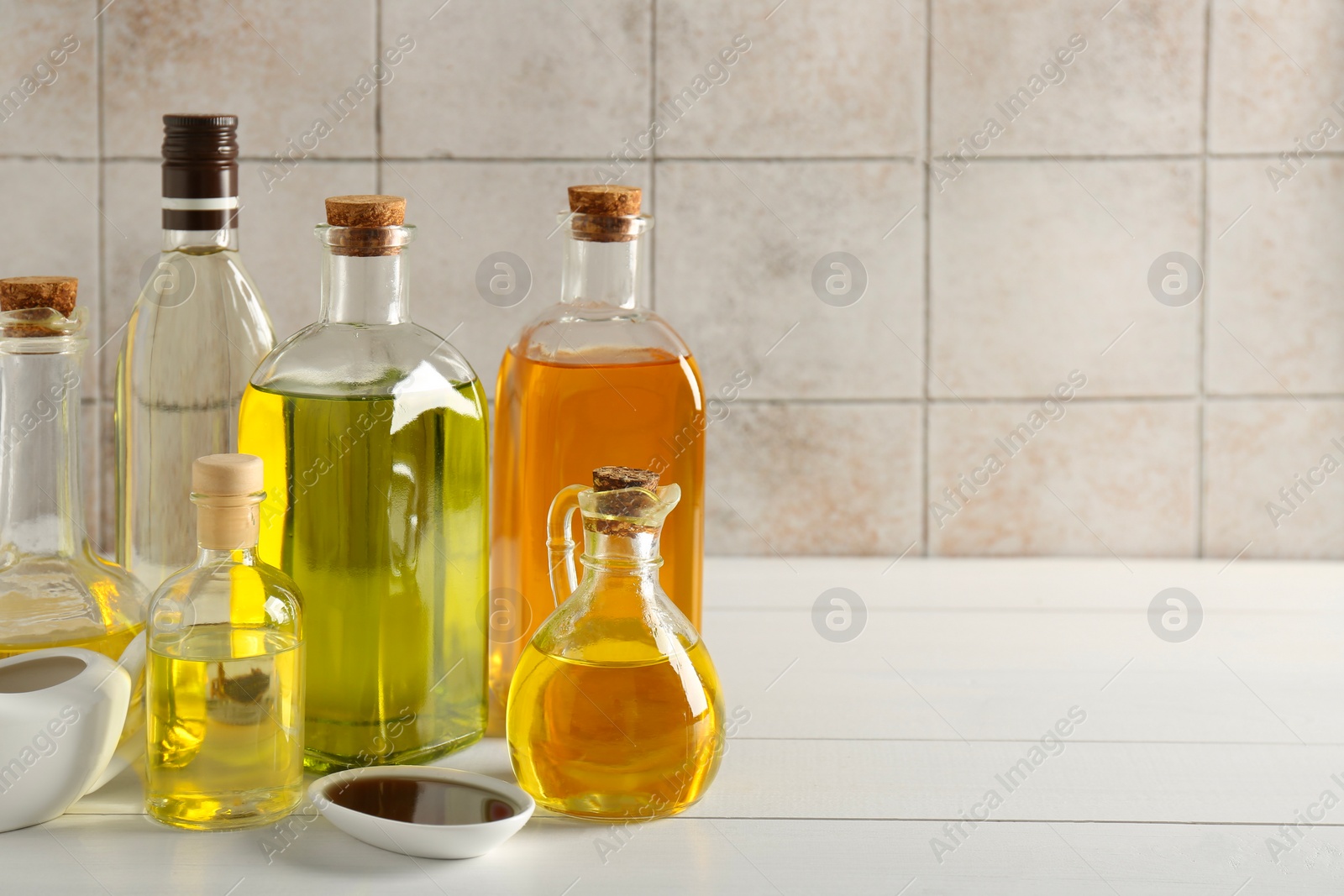 Photo of Vegetable fats. Different oils in glass bottles and dishware on white wooden table against tiled wall, space for text