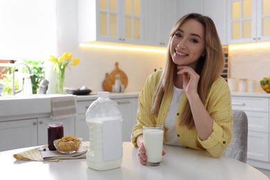 Young woman with gallon bottle of milk, glass and breakfast cereal at white table in kitchen