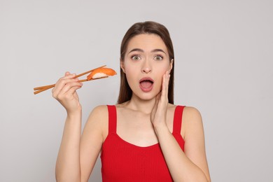 Emotional young woman holding sushi with chopsticks on light background