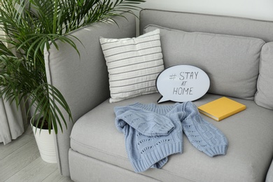 Photo of Book, sweater and speech bubble with hashtag STAY AT HOME on sofa indoors. Message to promote self-isolation during COVID‑19 pandemic