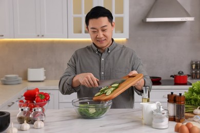 Photo of Cooking process. Man adding cut cucumber into bowl at countertop in kitchen