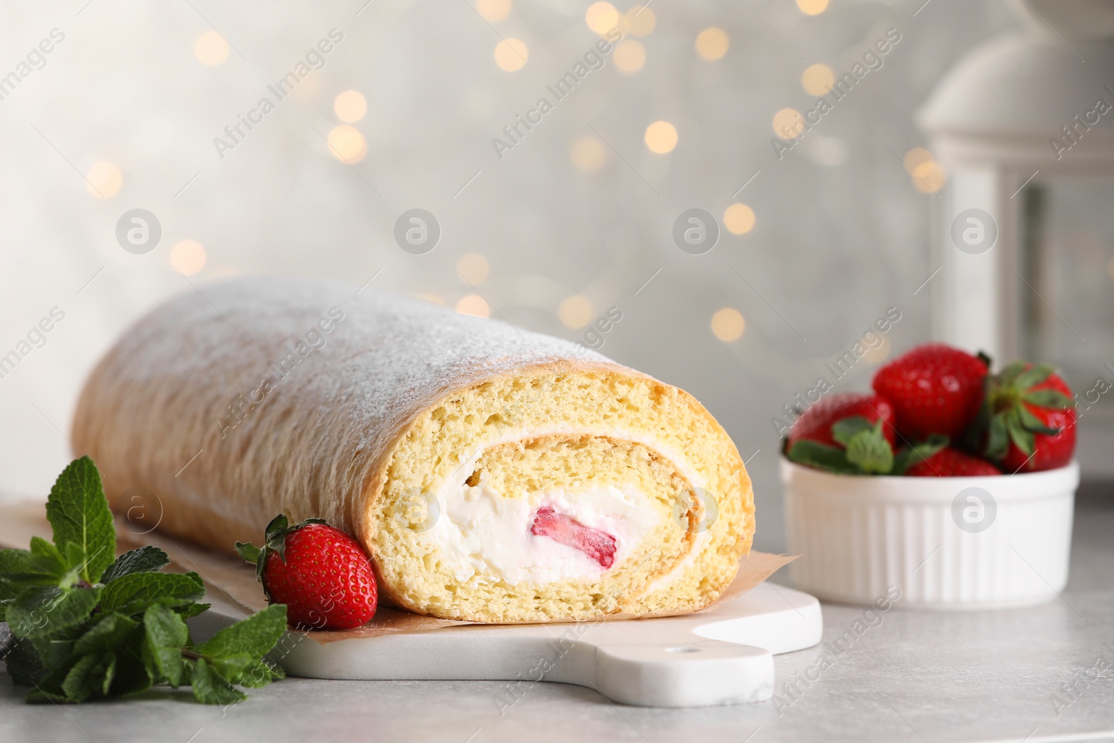 Photo of Delicious sponge cake roll with strawberries and cream on light grey table against blurred lights, space for text