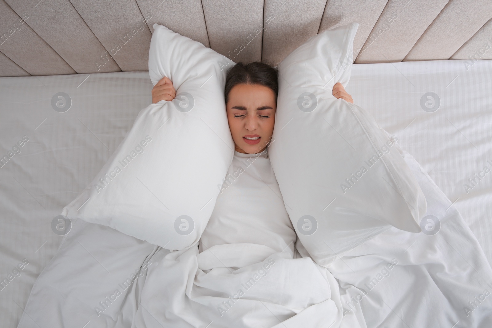 Photo of Unhappy young woman covering ears with pillows in bed at home, top view. Noisy neighbours