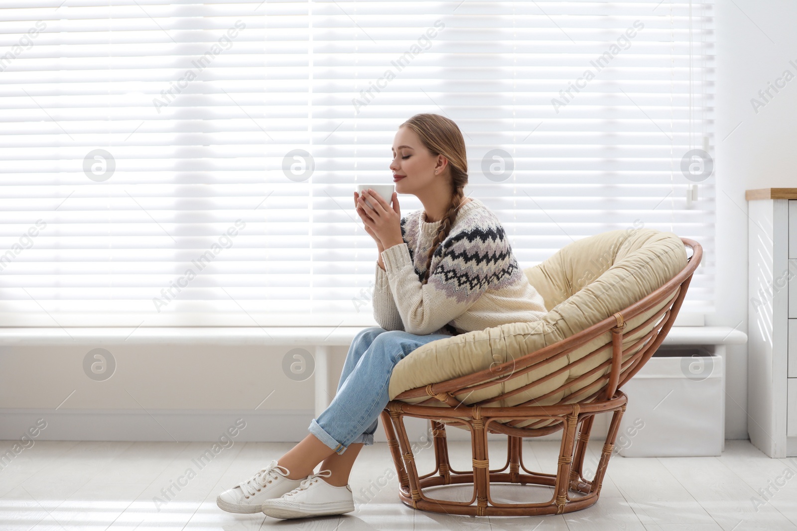 Photo of Young woman with hot drink in papasan chair near window at home