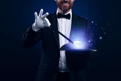 Image of Smiling magician showing trick with wand and top hat on dark blue background, closeup