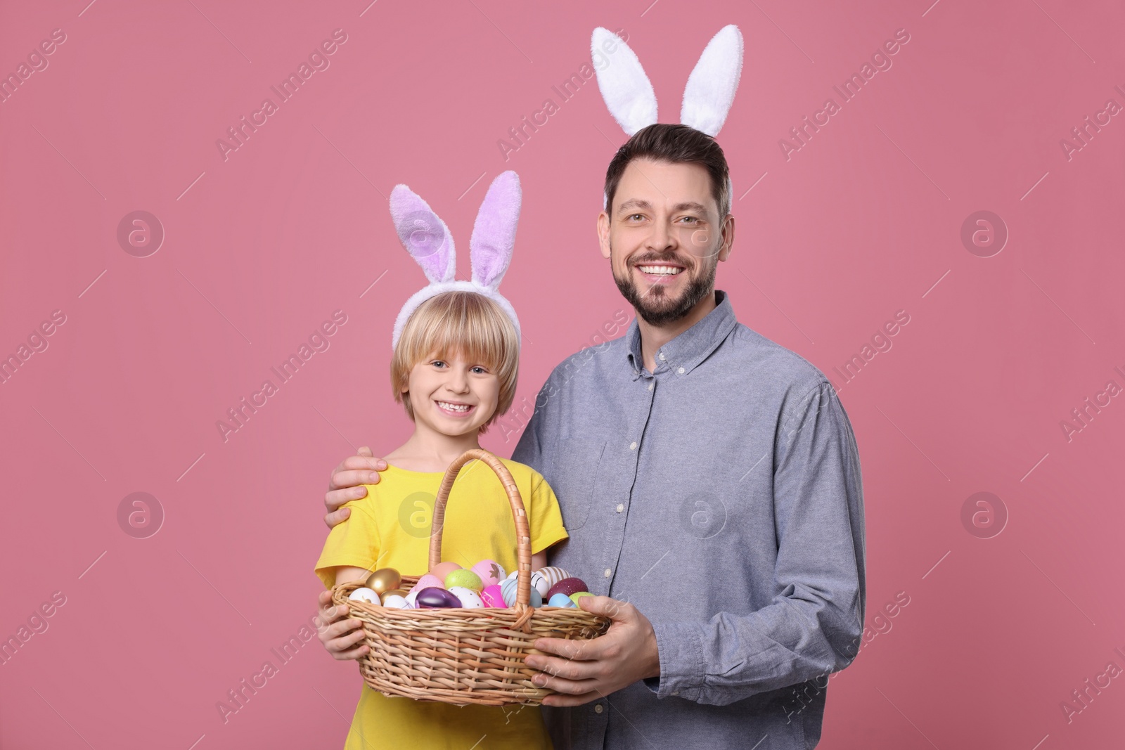 Photo of Father and son in bunny ears headbands with wicker basket of painted Easter eggs on pink background