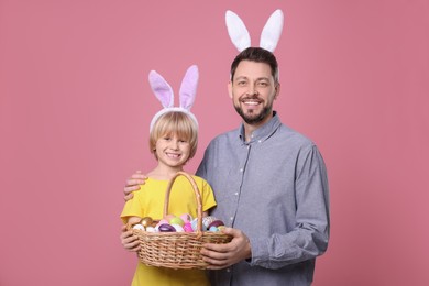 Photo of Father and son in bunny ears headbands with wicker basket of painted Easter eggs on pink background