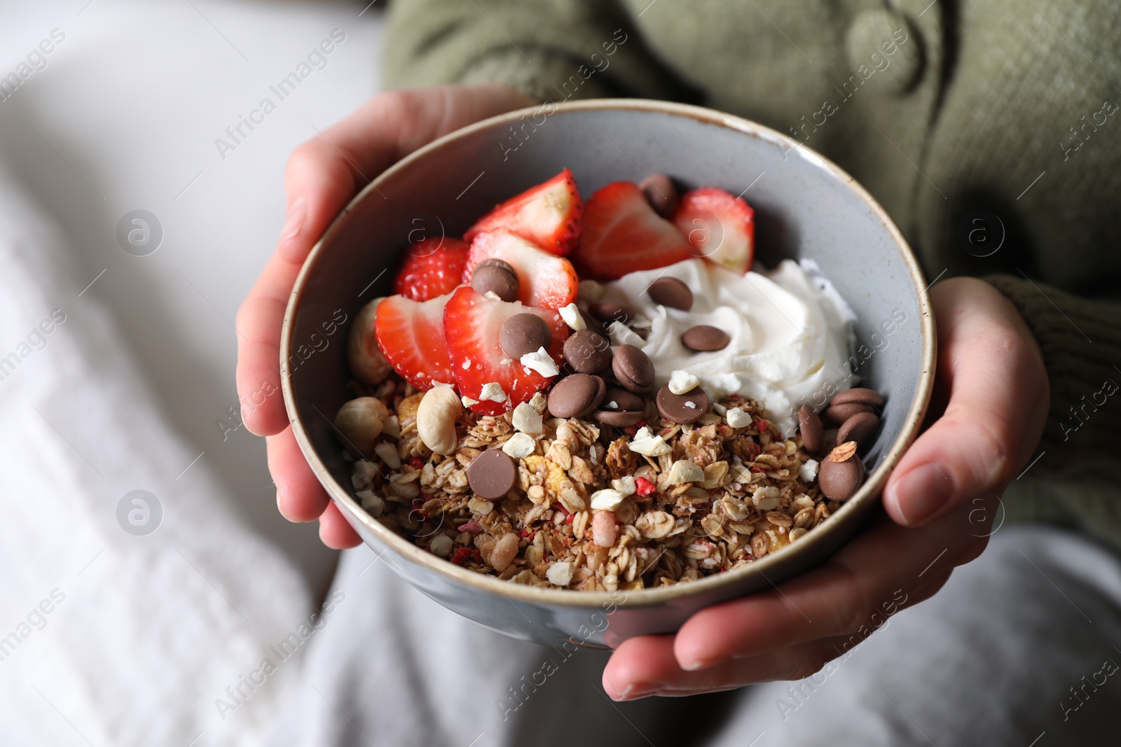 Photo of Woman holding bowl of tasty granola with chocolate chips, strawberries and yogurt indoors, closeup