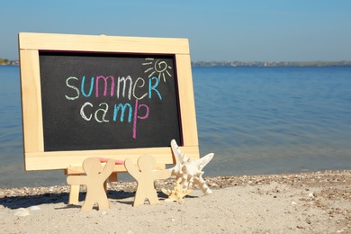 Photo of Small chalkboard with text SUMMER CAMP, wooden people and starfish on beach near river. Space for text