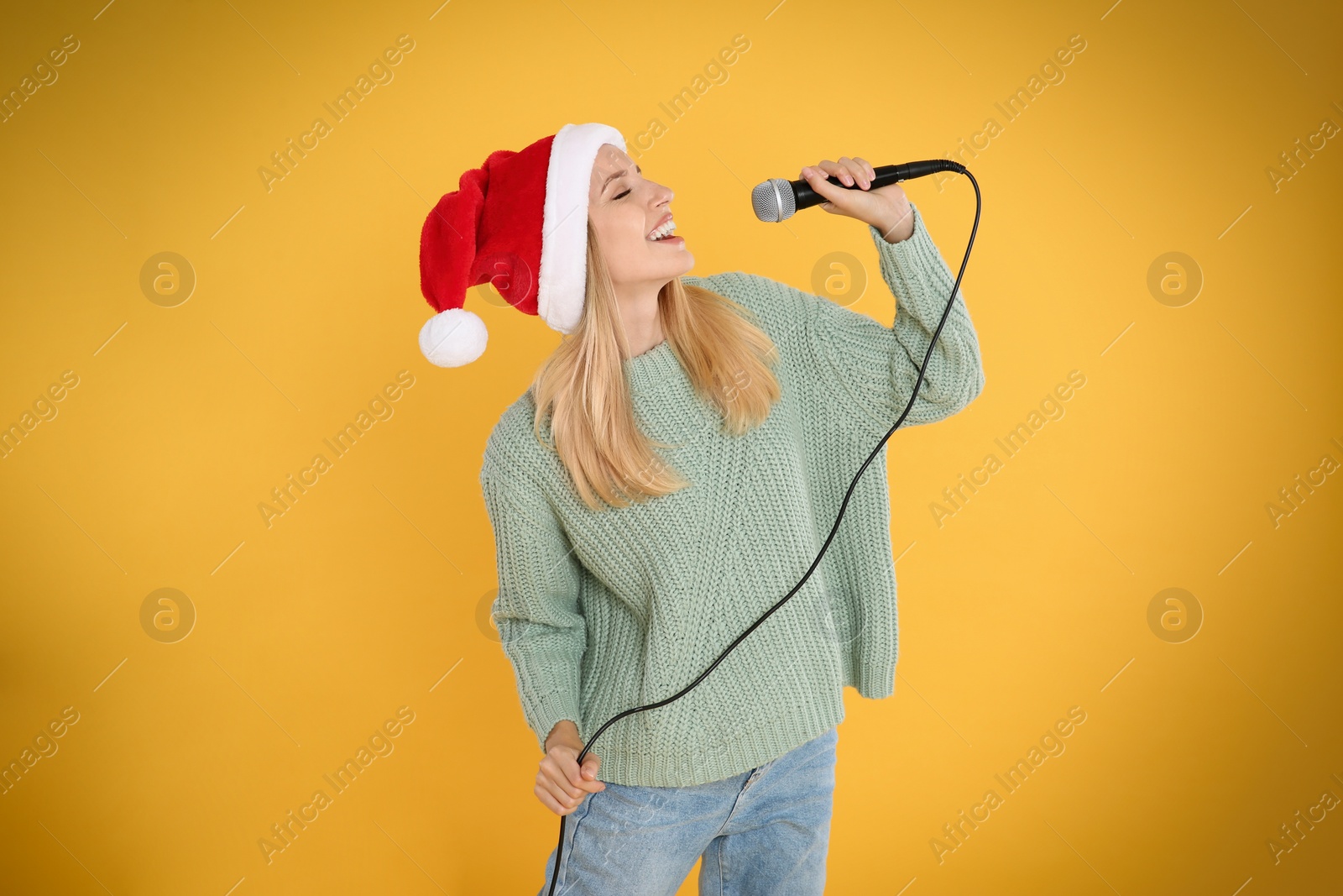 Photo of Happy woman in Santa Claus hat singing with microphone on yellow background. Christmas music