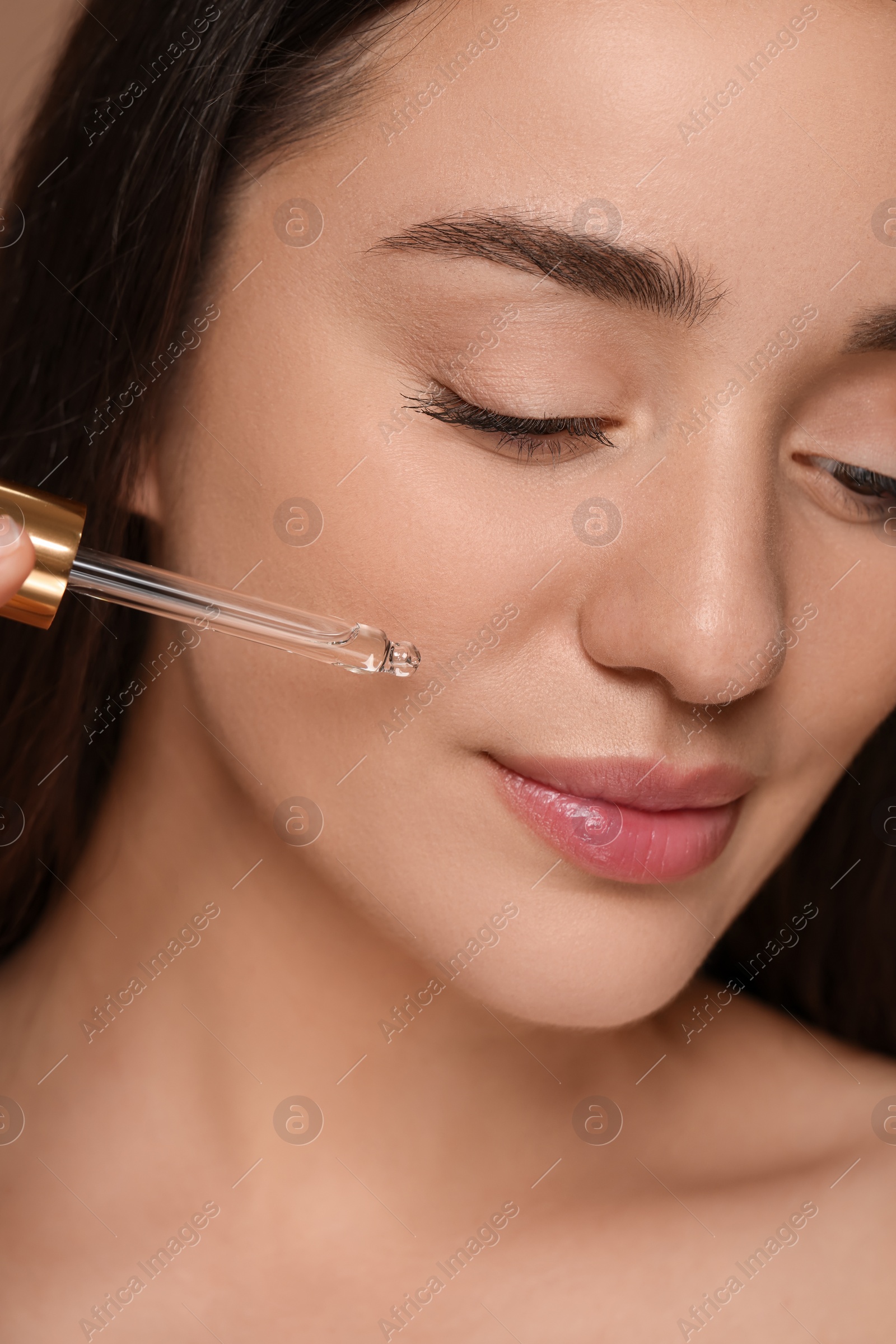 Photo of Beautiful woman applying essential oil onto face on brown background, closeup