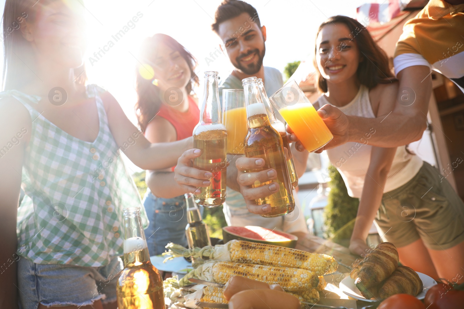 Photo of Happy friends toasting with beer and juice outdoors at table. Camping season