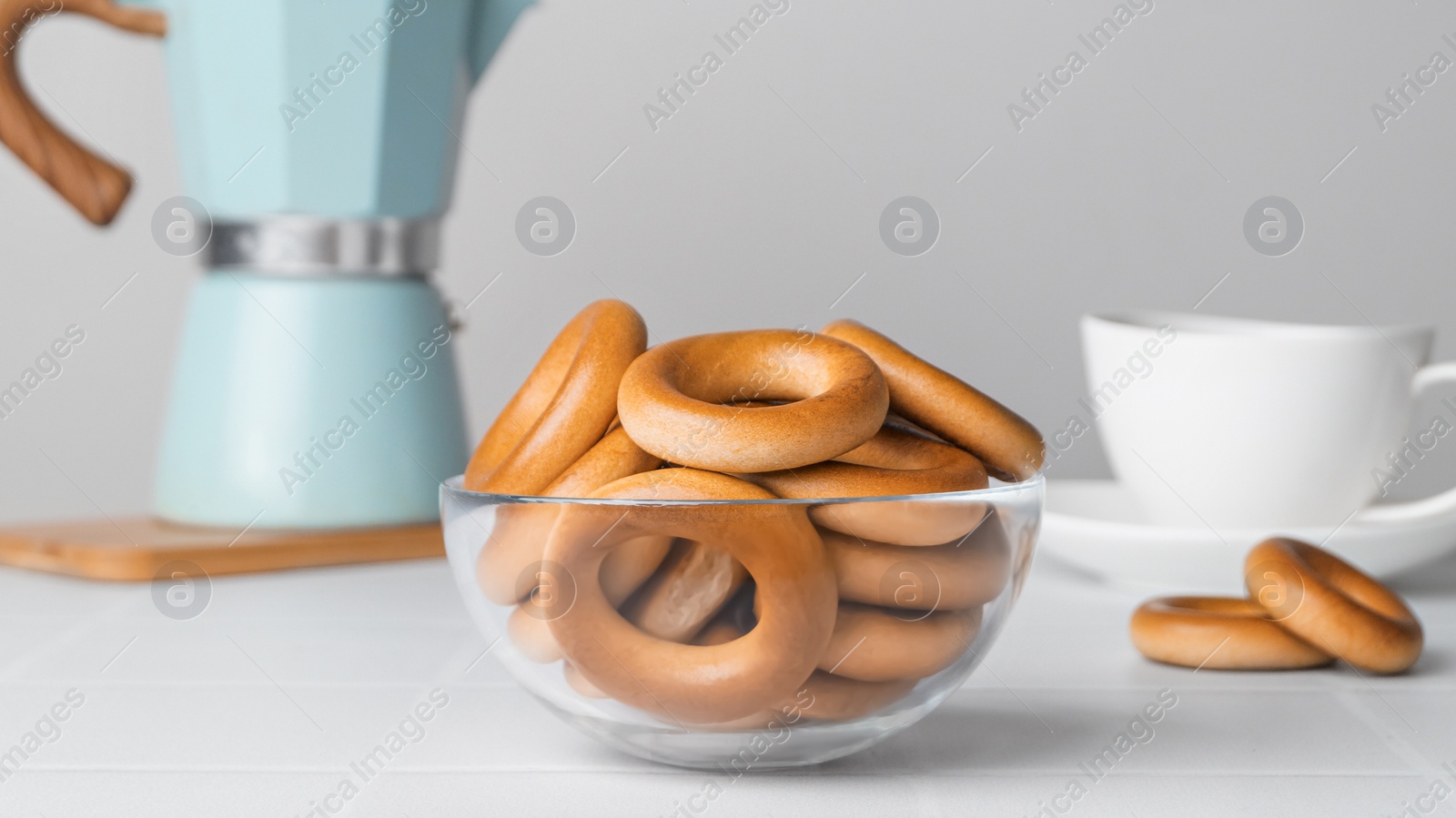 Photo of Bowl with tasty dry bagels (sushki) and cup of hot drink on white tiled table